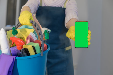 a woman holds a phone with a green screen and a plastic bucket with detergents, brushes, sponges and rubber gloves for cleaning.の写真素材
