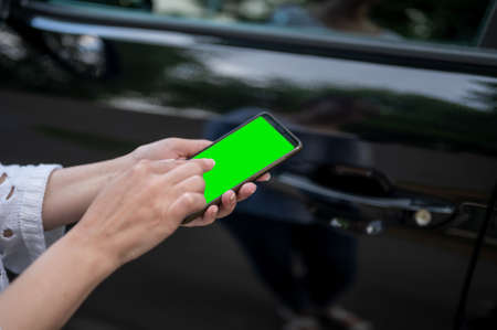 A woman holds a phone with a green screen in her hands against the background of a black car. a business woman stands with a phone by the car.の写真素材
