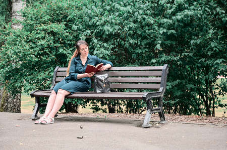Side view of a pleased brunette woman sitting on a bench and reading a book in the park.の写真素材