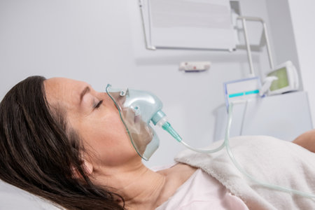 An adult woman lies on a hospital bed in an oxygen mask and undergoes treatment.