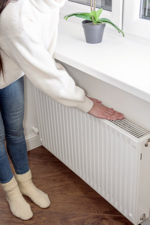 woman warms her hands. a woman standing next to a radiator in front of a windowの写真素材
