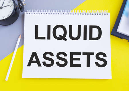 Conceptual hand writing showing Liquid Assets. Business photo text Cash and Bank Balances Market Liquidity Deferred Stock Open notebook squared page black marker computer wooden background.の写真素材