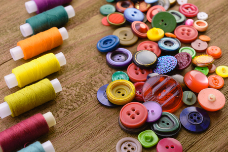 macro handful of multi-colored buttons for clothes near multi-colored threads on reels for sewing. wooden backgroundの写真素材