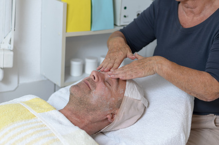 Senior man lying on bed at beauty center while receiving facial treatment through the application of hydrating or regeneration creamの写真素材