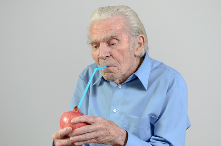 Conceptual image of a senior man holding a whole pomegranate  drinking the fresh pomegranate juice through a straw in a healthy diet and nutrition conceptの写真素材