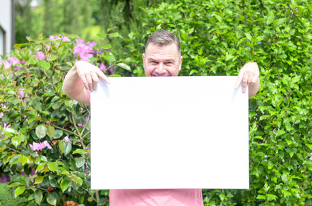 Young cheerful man smiling while holding and showing a blank whiteboard in a green garden with flowering shrubs in a sunny day of spring or summerの写真素材