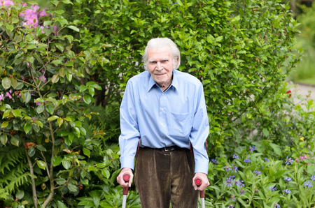 Elderly man with casual clothes using forearm crutches as a mobility aid to walk on the cobblestones of a footpath in a green park or yard full lengthの写真素材