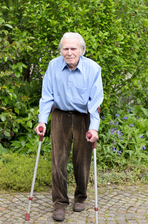 Elderly man with casual clothes using forearm crutches as a mobility aid to walk on the cobblestones of a footpath in a green park or yard full lengthの写真素材