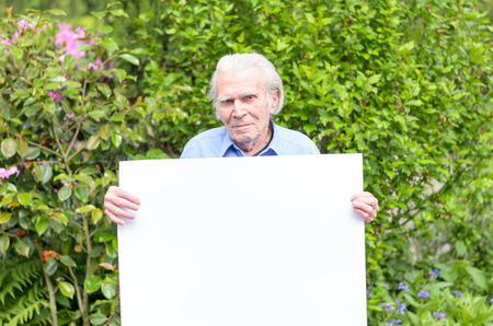 Serene elderly man smiling and showing a handheld blank whiteboard for advertising a message in front of a green flowering shrub behind himの写真素材