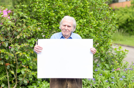 Serene elderly man smiling and showing a handheld blank whiteboard for advertising a message in front of a green flowering shrub behind himの写真素材