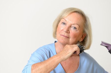 Senior woman blow drying her blond hair curling under it with a brush head and shoulders on a neutral gray backgroundの写真素材
