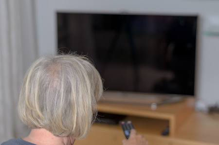 Rear View of a White Haired Middle Aged Woman Sitting at the Living Area While Controlling the TV Using a Remote Control Device.の写真素材