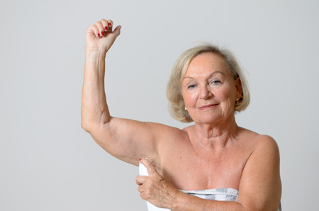 Close up Middle Aged Blond Lady Applying Deodorant on Armpit After Shower While Looking at the Camera Against Light Gray Wall.の写真素材