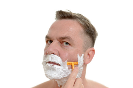Middleaged man shaving his beard with a razor and lather from shaving cream covering his face isolated on whiteの写真素材