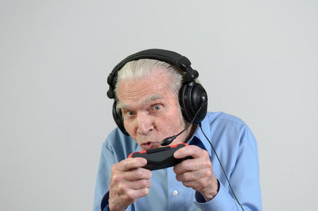 Funny elderly man or grandfather using black headset with headphones and microphone while playing a video game on console portrait with copyspace on grayの写真素材