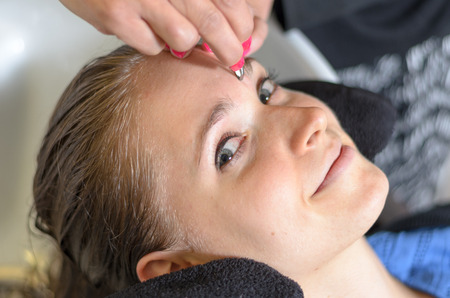 Beautician trimming a young girls eyebrows with a pair of tweezers as she relaxes with her head in the basin closeup of her faceの写真素材