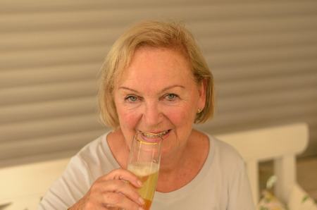 Attractive senior lady drinking cold champagne form an elegant flute raising her glass to her lips,...