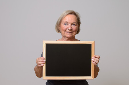 Happy senior woman with blond hair showing a blank small blackboard while smiling at camera, portrait with copy space on greyの写真素材