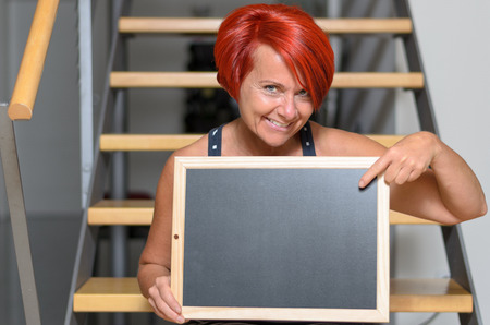 Close up Adult Redhead Woman Holding Small Chalkboard with Copy Space While Sitting on the Stairs and Smiling at the Camera.の写真素材