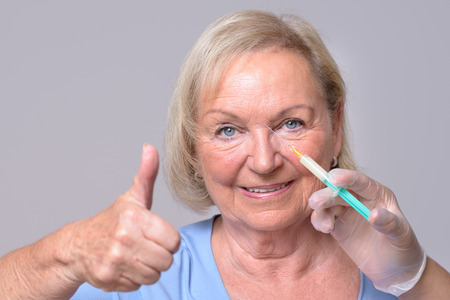 Close up Satisfied Middle Aged Woman with Injection on her Face, Showing Thumbs Up Hand Sign and Smiling at the Camera. Isolated on Gray Background.の写真素材