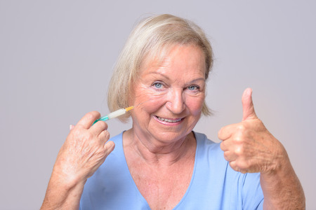 Close up Happy Middle Aged Woman Holding Injection, Showing Thumbs Up Hand Sign and Smiling at the Camera. Isolated on Gray Background.の写真素材