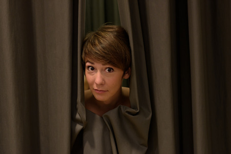 Attractive woman peeking out from drawn curtains framing her face with a quizzical expression
