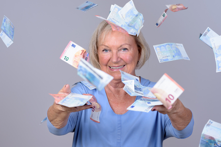 Excited senior lady celebrating a windfall or win throwing Euro banknotes into the air with a happy smile , over a grey backgroundの写真素材
