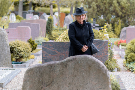Senior lady in mourning at a graveside standing dressed in full black paying her respects to a departed loved one in a large graveyardの写真素材