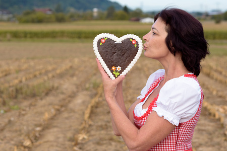 Attractive young Bavarian woman wearing a traditional red dirndl standing in a rural field kissing a heart shaped decorative biscuit symbolic of love and romance and Valentines Dayの写真素材