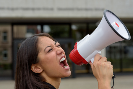 Angry young woman yelling over a megaphone or bullhorn as she participates in a street demonstration or protest airing her grievances, close up side view of her faceの写真素材