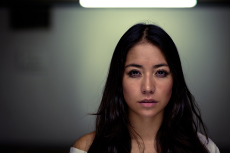 Head and shoulders portrait of a pretty serene dark haired young woman looking directly at the camera under the glow of an overhead light, with copyspaceの写真素材