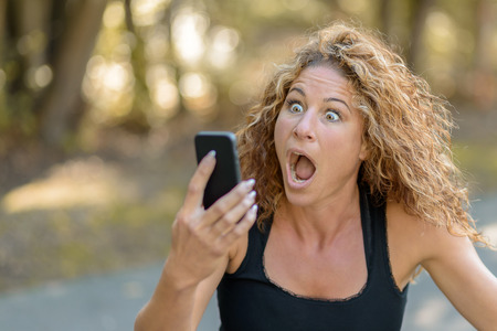 Attractive young woman with gorgeous curly long hair reacting in horror to a text message on her mobile phone staring at it with an aghast expression and mouth openの写真素材