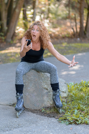 Horrified attractive sporty young woman staring at her mobile phone with a shocked expression as she sits on a rock at the side of road in her roller bladesの写真素材