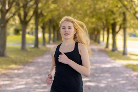 young blond woman jogging on a path with big green trees while looking at the cameraの写真素材