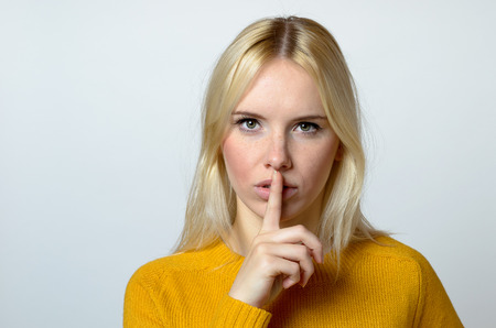 Close up Pretty Blond Woman Showing Shushing Gesture While Staring at the Camera Against Gray Background.の写真素材