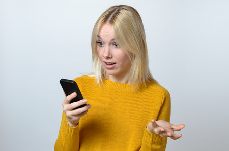 Blond Young Woman Showing Surprise Expression While Looking at her Mobile Phone Against White Background.の写真素材