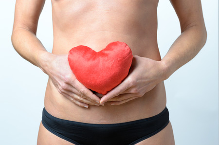 middle aged woman holding a pillow which looks like a red heart in front of her belly button, close upの写真素材