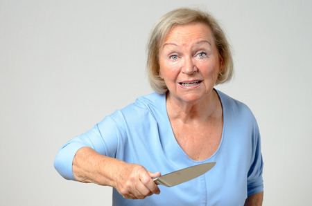 Emotional elderly woman brandishing a kitchen knife in her hand with a look of perplexed horror and shock, over greyの写真素材