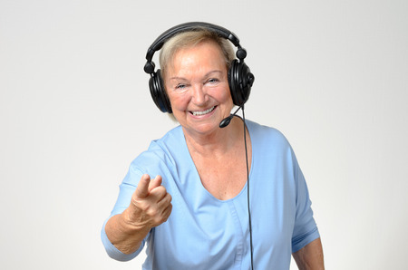 Excited senior woman listening to music on a set of stereo headphones smiling with enjoyment and pointing at the camera, over greyの写真素材