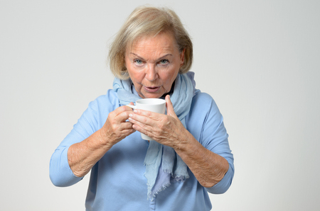 Elderly woman in blue winter fashion standing enjoying a hot cup of coffee to warm herself, upper body over greyの写真素材