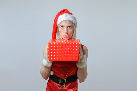 Sulking woman wearing a festive red Santa hat standing holding a decorative red polka dot wrapped Christmas gift in her hand as she celebrates the holiday seasonの写真素材