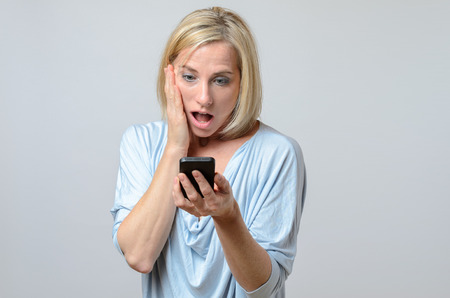 Surprised middle-aged woman with blue eyes and a shocked facial expression looking at the screen of her mobile phone, portrait with copy space on grayの写真素材