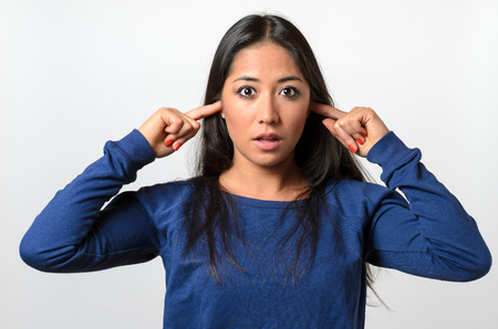 Shocked young woman blocking her ears with her fingers as she stares wide-eyed at the camera with an appalled expressionの写真素材