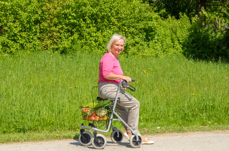 Smiling elderly woman wearing pink blouse while seated in walker with wheels on park pathの写真素材