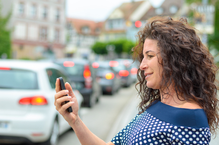 Attractive woman reading a text message on her mobile phone with a serious expression as she stands on a busy urban street with trafficの写真素材