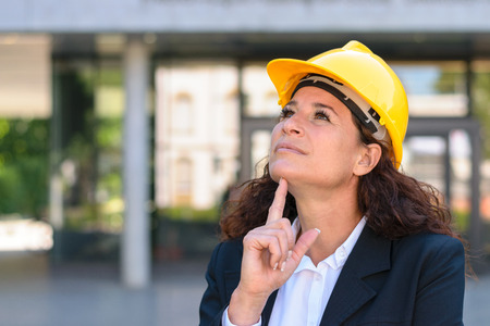 Thoughtful young female architect or building inspector standing staring up into the air with a pensive expression in a yellow hardhatの写真素材