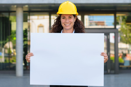 Attractive young female architect or structural engineer wearing a hardhat standing in front of a glass fronted building in town holding a blank white signの写真素材