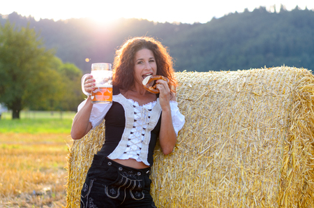 Attractive woman enjoying the Oktoberfest leaning against a round hay bale in a field drinking beer and eating a pretzelの写真素材