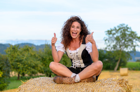 Happy exited woman in a Bavarian outfit sitting on a hay bale laughing and cheering while giving a double thumbs up signの写真素材