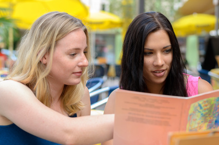 Pair of beautiful young adult women looking at book or menu together at outdoor cafeの写真素材